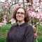 A person with shoulder-length curly hair and glasses standing in front of a tree covered in pink flowers